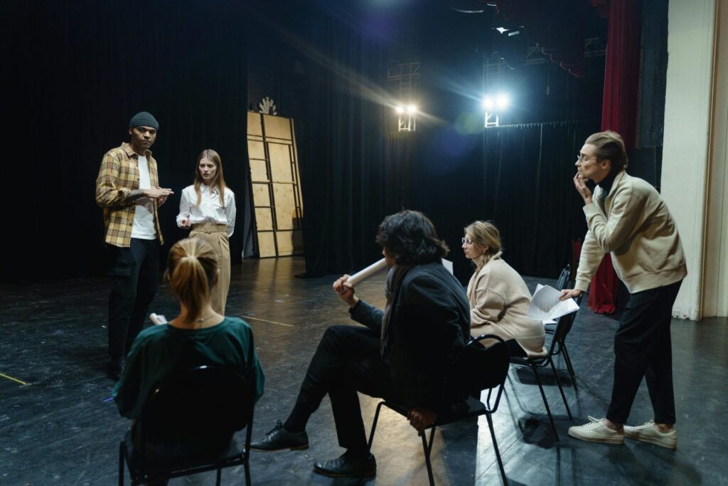 Jugendliche nehmen an einem Workshop im First Steps Schauspiel teil und sitzen auf Stühlen in einem Theater.